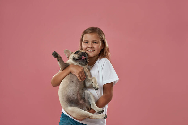 Joyful little blonde girl holding French bulldog - Fotografie, Obrázek