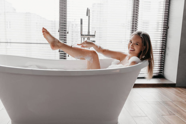 Happy lady ontspannen in bad en scheren benen met behulp van scheermes voor ontharing, kijken en glimlachen op de camera - Foto, afbeelding