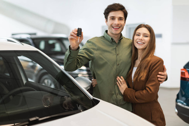 Joyful young Caucasian couple holding new car key, smiling at camera in modern dealership store - Foto, imagen