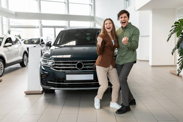 Первая семейная машина. Счастливая молодая кавказская пара трясет кулаками, делая ДА жест после покупки нового автомобиля в автосалоне - Фото, изображение