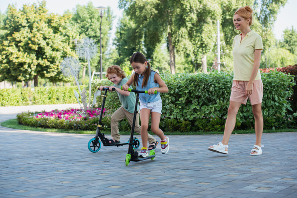 femme heureuse marchant près des enfants équitation coup de pied scooters dans le parc - Photo, image