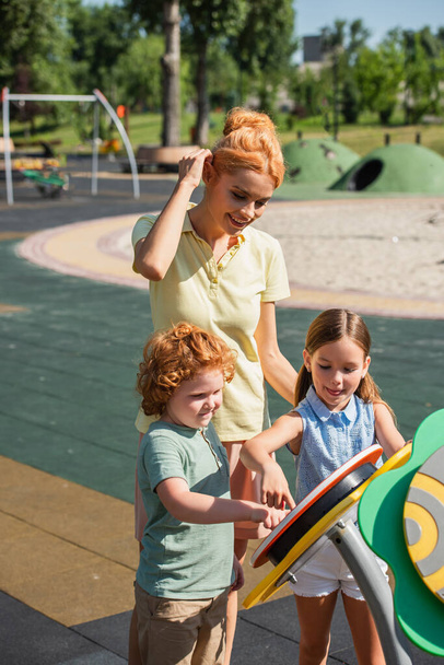 uśmiechnięta kobieta dopasowanie włosów w pobliżu dzieci zabawy na placu zabaw - Zdjęcie, obraz
