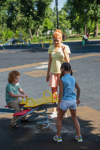 potěšená žena stojí v blízkosti dětí a houpačka v parku - Fotografie, Obrázek