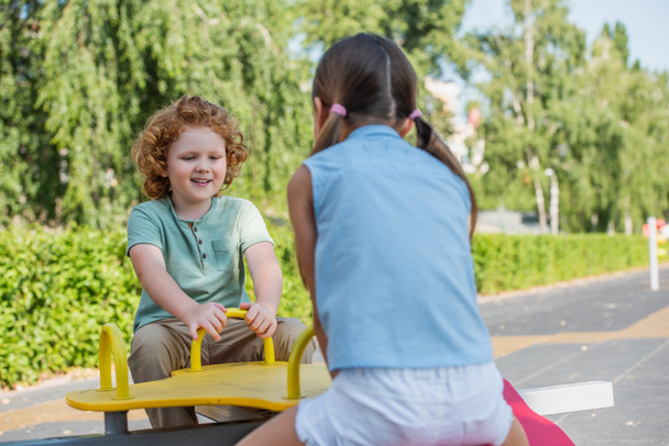 веселый мальчик катается на качелях с сестрой в летнем парке - Фото, изображение
