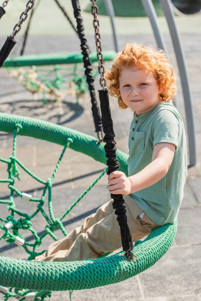 curly boy looking at camera while having fun in hammock in amusement park - Photo, Image