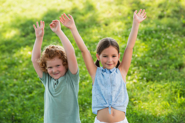 happy brother and sister looking at camera while standing with raised hands outdoors - Фото, изображение