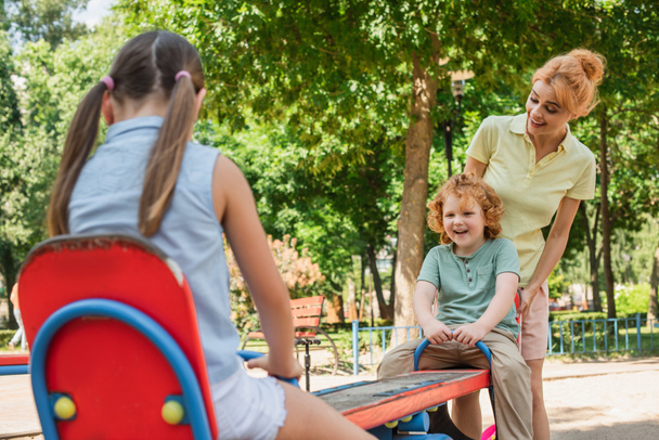 feliz mamá de pie cerca de los niños montando balancín en el patio de recreo - Foto, Imagen