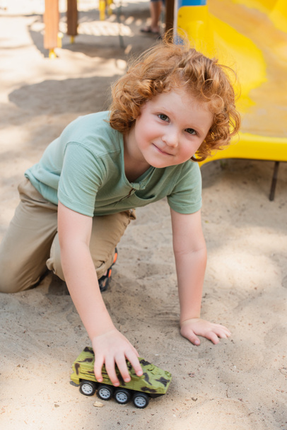 кудрявый мальчик смотрит в камеру, играя на песке с игрушечным бронированным автомобилем - Фото, изображение