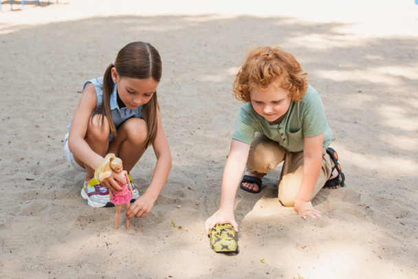 άποψη πλήρους μήκους των παιδιών που παίζουν με κούκλα και θωρακισμένο όχημα στην άμμο σε εξωτερικούς χώρους - Φωτογραφία, εικόνα