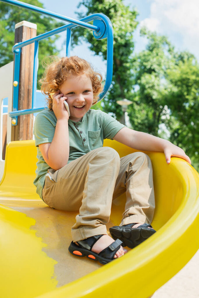 веселый мальчик разговаривает на смартфоне на слайде в парке развлечений - Фото, изображение