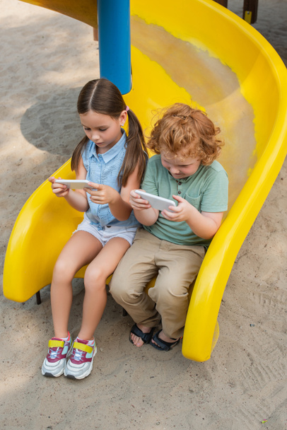 korkea kulma näkymä lapset käyttävät älypuhelimia istuessaan liukumäki - Valokuva, kuva