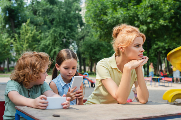 Düşünceli ve üzgün bir kadın parktaki akıllı telefonlarla oynayan çocukların yanından uzağa bakıyor. - Fotoğraf, Görsel
