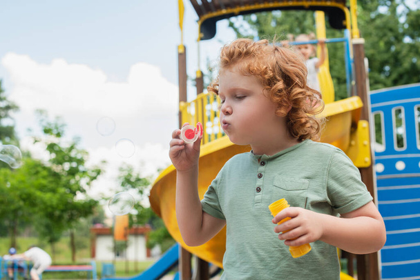 curly boy blowing soap bubbles on playground - Fotoğraf, Görsel