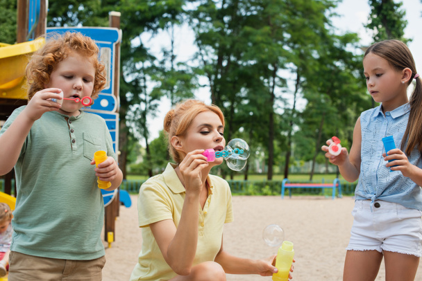 vrouw blazen zeepbellen met kinderen op speelplaats - Foto, afbeelding