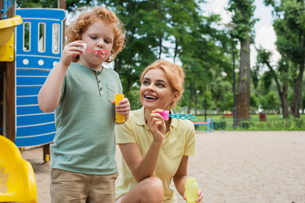 Rotschopf pustet Seifenblasen neben fröhlicher Mutter auf Spielplatz - Foto, Bild