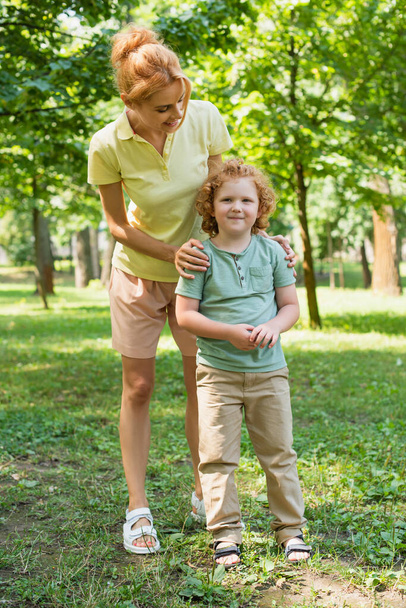 pełna długość widok kobieta obejmując ramiona syna podczas stojąc na trawniku w parku - Zdjęcie, obraz