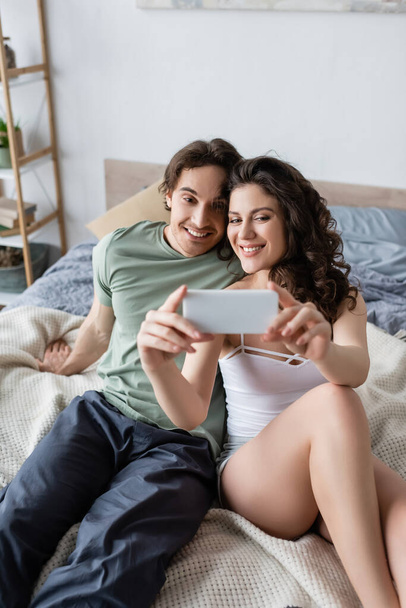 mujer rizada y hombre feliz tomando selfie en el dormitorio - Foto, imagen