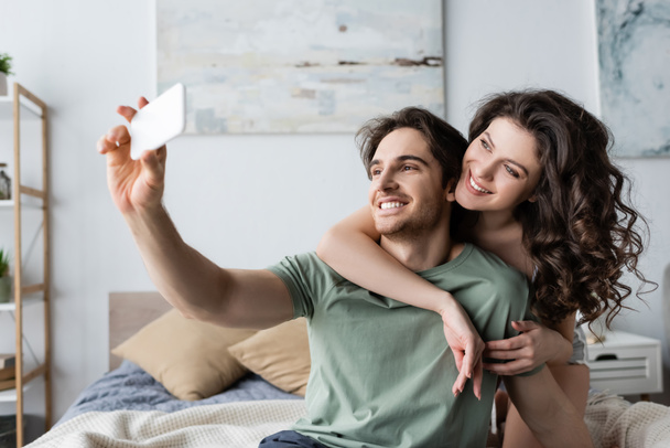 feliz joven pareja tomando selfie en dormitorio
 - Foto, Imagen
