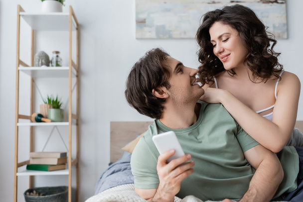 jonge man met smartphone en kijken naar vriendin in de slaapkamer - Foto, afbeelding