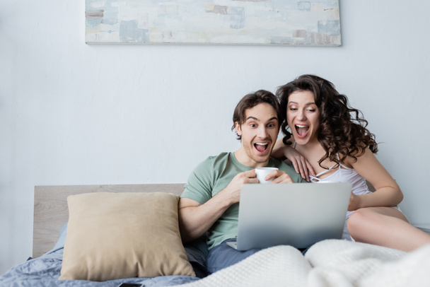 excited young couple looking at laptop in bedroom - 写真・画像