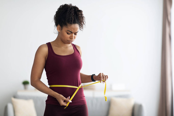 Unhappy african american woman in sportswear measuring waist - Foto, afbeelding