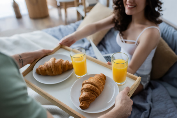 обрезанный вид на татуированного человека, держащего поднос с вкусным завтраком возле счастливой подружки в постели - Фото, изображение