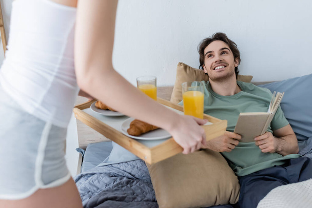 blurred woman holding tray with tasty breakfast near smiling boyfriend with book - Fotografie, Obrázek