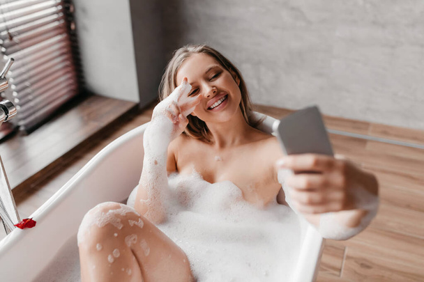 Mujer alegre tumbada en un baño espumoso y tomando selfie en el teléfono inteligente, relajándose y disfrutando de la rutina matutina de belleza - Foto, imagen