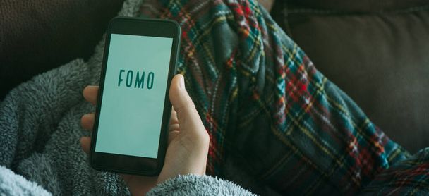 closeup of a young man in the sofa, wearing pajamas and house robe, holding his smartphone, that reads the text fomo, for fear of missing out, in his hand, in a panoramic format to use as web banner - Photo, Image