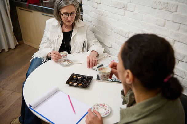 Two diverse people of two generations sitting at a table and enjoying a discussion while drinking coffee in a cozy fashion design studio - 写真・画像