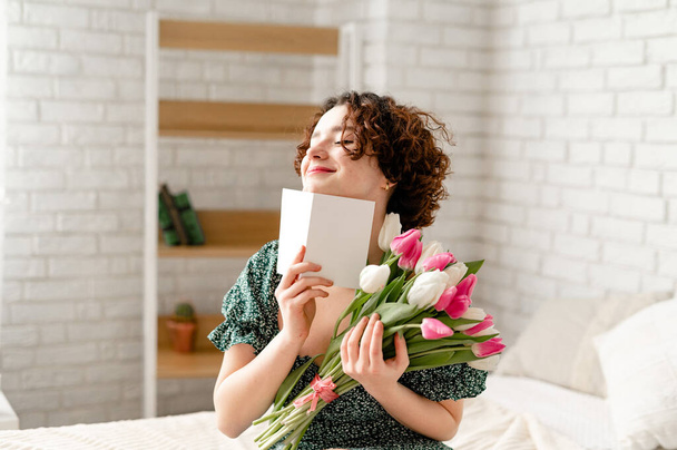 Όμορφη έκπληξη μελαχρινή κοπέλα με μια κάρτα που κρατά ένα μπουκέτο τουλίπες και κάθεται στο κρεβάτι - Φωτογραφία, εικόνα