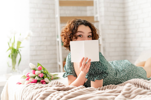 a brunette girl with curls lies on the bed and covers her face with a card. next to a bouquet of tulips on the bed - Foto, Imagem