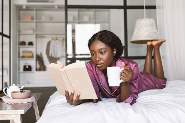 African American Woman Reading Book Drinking Coffee In Bedroom - Foto, afbeelding