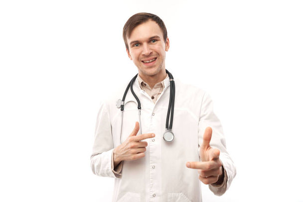 Portrait of friendly positive young male doctor in white medical coat chooses you, points finger at camera isolated on white backgroun - Foto, Imagen