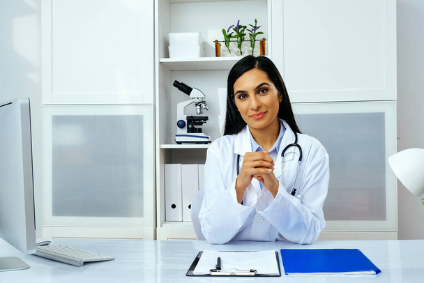 Retrato de una hermosa doctora joven en uniforme blanco de bata de laboratorio sentada en su escritorio en su oficina moderna - Foto, imagen