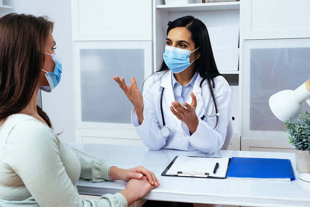 Doctor and patient in face masks discussing health problems while sitting at the table in medical center  - 写真・画像
