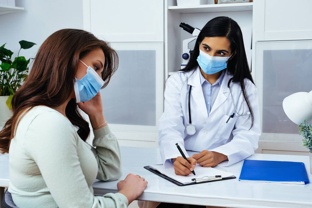 Lekarz i pacjent w maskach do twarzy omawiając problemy zdrowotne podczas siedzenia przy stole w centrum medycznym  - Zdjęcie, obraz
