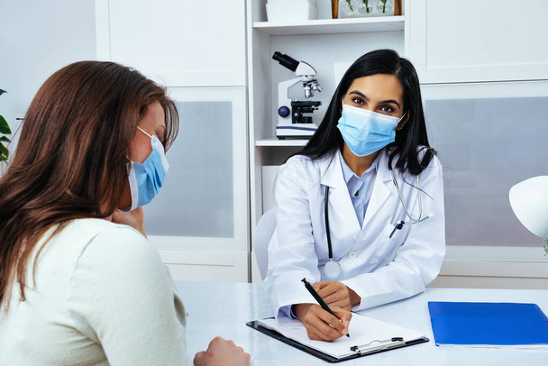 Врач и пациент в масках для лица обсуждают проблемы со здоровьем сидя за столом в медицинском центре  - Фото, изображение