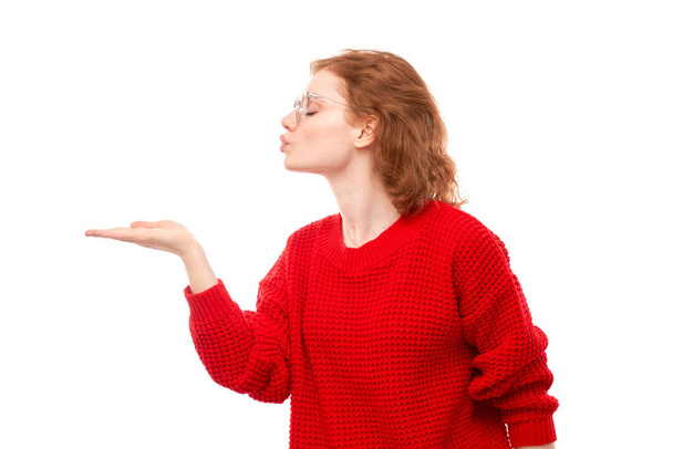 Attractive redhead girl promoter in red sweater holding something in hand demonstrating empty space with excited face isolated on white background - Photo, Image