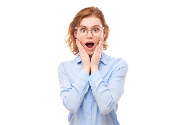 Σοκαρισμένο κορίτσι τζίντζερ πρόσωπο σε επιχειρηματικό πουκάμισο φαίνεται έκπληκτος με ανοιχτό στόμα κρατώντας το κεφάλι με τα χέρια σε λευκό φόντο στούντιο - Φωτογραφία, εικόνα