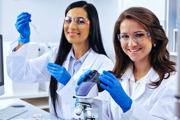 Две красивые молодые женщины-учёные, работающие в лаборатории с пробирками и микроскопом, проводят некоторые исследования  - Фото, изображение