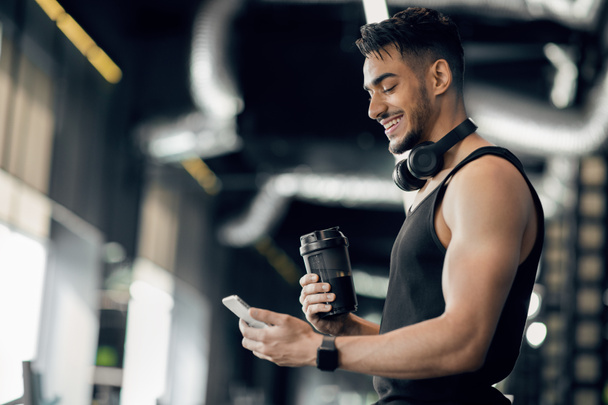 Εφαρμογή γυμναστικής. Χαμογελώντας Άραβας Sporty άνθρωπος χρησιμοποιώντας Smartphone, ενώ στέκεται στο γυμναστήριο - Φωτογραφία, εικόνα