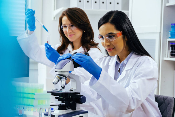 2人の美しい若い女性科学者が実験室で実験管と顕微鏡を使って研究をしています  - 写真・画像