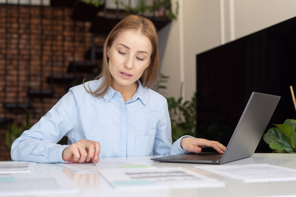 Portrait of focused woman with laptop carefully verifies financial report results. Home office concept. Process of utility bills payment online. Businesswoman checking information on paper - Photo, Image