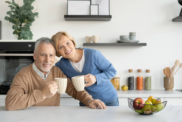 Portré boldog nyugdíjas házastársakról, akik poharakat tartanak és kamerába néznek. Reggeli kávét iszunk együtt. Otthon kényelem. Örök szerelem. Szabadidő hétvége - Fotó, kép