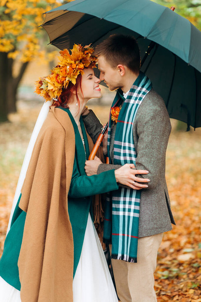 Eine schöne rothaarige Braut in einem Kranz aus Herbstblättern berührt die Nase des Bräutigams - Foto, Bild