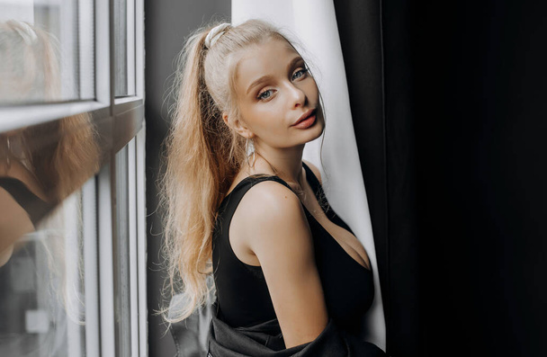 Stylish blonde in fashionable black clothes posing near the window. - Foto, Bild