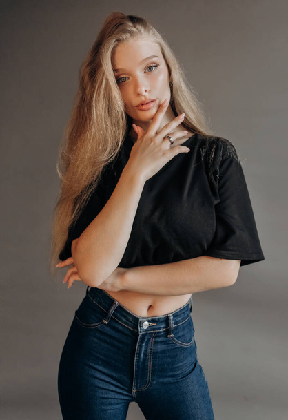 Pretty girl with blond hair dressed in a short top and jeans posing in the studio on a gray background - Zdjęcie, obraz