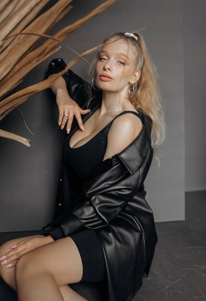 Amazing blonde model dressed in a black fashionable suit sitting on a gray background. - 写真・画像