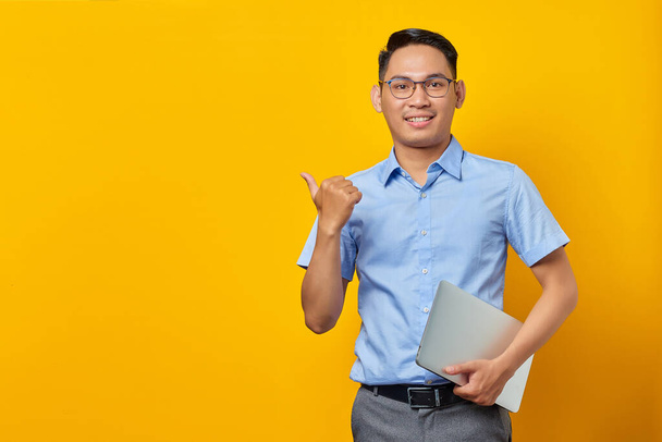 Portrait of smiling handsome Asian man in glasses holding laptop and pointing fingers at copy space isolated on yellow background. businessman and entrepreneur concept - Φωτογραφία, εικόνα